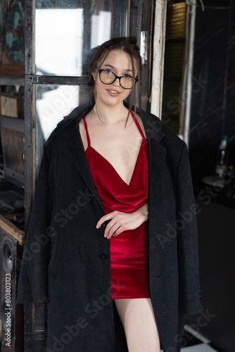young beautiful woman posing in a short red dress in the studio wearing glasses and a black coat