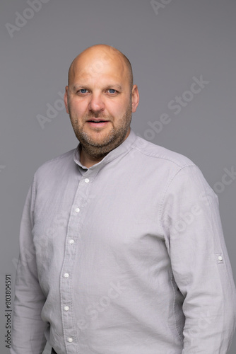 A studio portrait of a bald man in a light grey shirt, looking directly at the camera with a slight smile.