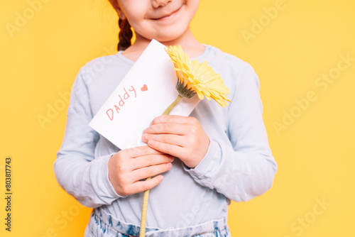 Child holding drawn card and flower for Father's Day holiday in front of yellow background