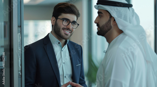 A businessman wearing a suit and glasses is talking to an Emirati guy in a thobe, both of them with their hands on the door. photo