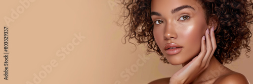 A mixed race woman with curly hair is posing for a portrait against a beige background. She has beautiful eyebrows and soft pink lips.