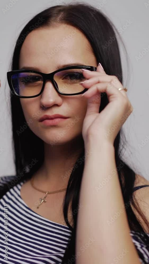 Serious young girl tries on fashionable eyeglasses in black rim over the light background. Attractive woman puts on new eyewear. Close-up Vertical video Serious young girl tries on fashionable eyeglasses in black rim over the light background. Attractive woman puts on new eyewear. Close-up Vertical video