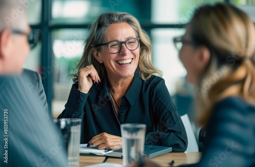 Happy business team having fun laughing together at the meeting table, a smiling middle aged woman leader talking with colleagues in the office room