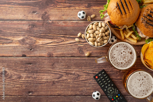 Football frenzy fare. Top view of snack assortment: crispy tortilla chips, savory pistachios, hamburgers, ice-cold beer, soccer balls, remote control, on wooden table with empty space for text