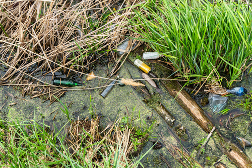 Polluted pond water with scattered litter among green grass and dead reeds, including bottles, plastic, and miscellaneous debris close-up
