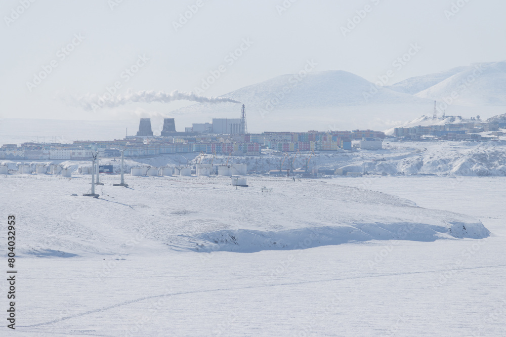 Winter Arctic landscape. View of the northern city. Anadyr, Chukotka, Russia. In the foreground are wind turbines and large fuel tanks at Cape Observation. Snowy tundra on the coast of a frozen river.