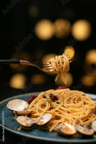 A hand holding a fork with spaghetti vongole on dark blue ceramic plate in black background and bokeh warm color light. Closed up.
