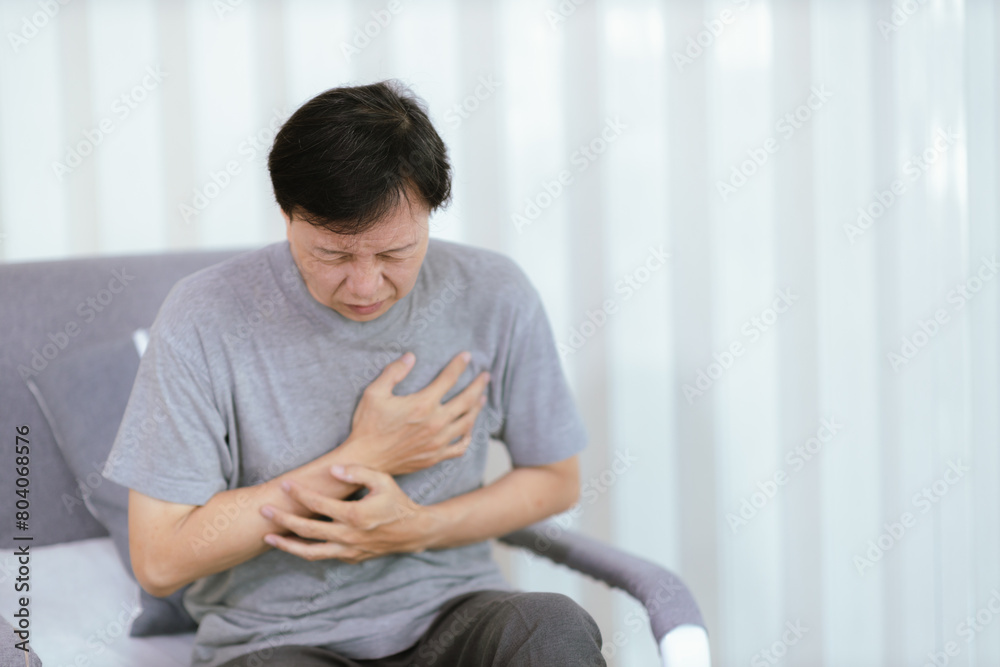 An elderly Asian man having a heart attack sits on a sofa in the living