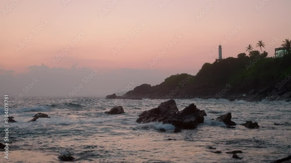 Sunset paints sky in soft pink hues by a hillside lighthouse. Waves crash against rocky shore. Peaceful scenery for relaxation, coastal ambience attracts nature lovers. Slow motion. Sunset paints sky in soft pink hues by a hillside lighthouse. Waves crash against rocky shore. Peaceful scenery for relaxation, coastal ambience attracts nature lovers. Slow motion.