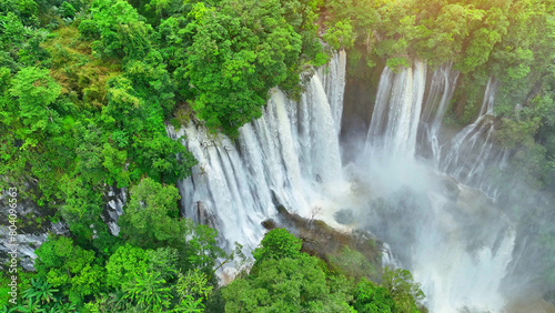 A mesmerizing hidden gem in the heart of the lush tropical rainforest. A grand waterfall cascades through the dense green canopy, captured by a drone's soaring lens. Primitive woods. Thailand. 