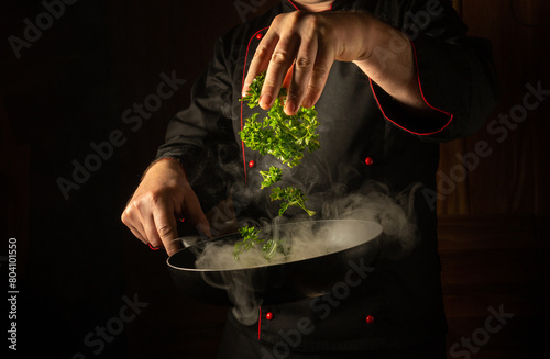 The chef adds fragrant parsley with his hand to a steaming hot frying pan with scrambled eggs. Low key concept of preparing breakfast in a hotel kitchen. Thai cuisine