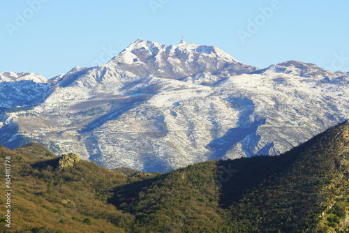 View of snow-capped Mount Lovcen against a blue sky and a fragment of forested Mount Vrmac below - a winter mountain landscape from Montenegro.
