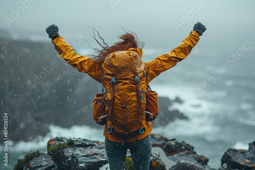 A person in a yellow jacket stands on a rocky cliff with arms raised in triumph amid foul weather photo