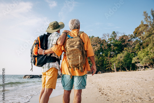 Low angle rear view of senior gay couple with backpacks standing at beach on vacation photo
