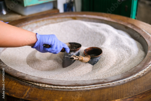 In a traditional Indonesian kopitiam, a hand in a blue glove stirs coffee grounds in fine sand using two small pots. The unique Turkish-style brewing method creates a rich aroma and flavor