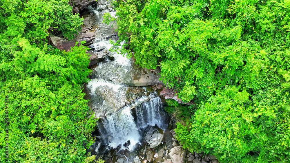 Aerial view of a stunning tropical waterfall, flowing down a towering cliff amidst a lush forest paradise. Thung Na Muang Waterfall, Pha Taem National Park, Ubon Ratchathani Province, Thailand.
