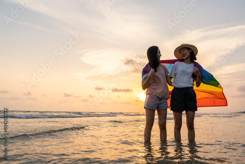 Young couple asian lesbian with pride movement LGBT holding rainbow flag for freedom. Demonstrate rights LGBTQ celebration pride Month lesbian Pride Symbol. Walking on the sand sea beach with sunset photo
