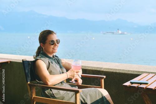 Outdoor portrait of happy relaxing woman, enjoying nice sunny day by the lake, holding glass of fresh beverage