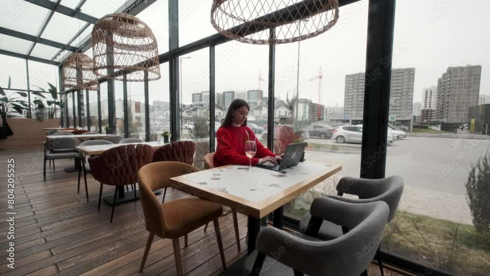 A beautiful young girl in a red jacket is sitting at a table in a restaurant. Working at a laptop. Remote work concept. A beautiful young girl in a red jacket is sitting at a table in a restaurant. Working at a laptop. Remote work concept.