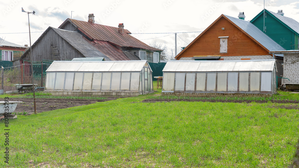 Green grass on the field behind the farmer's house, farm and agricultural production concept. Two greenhouses for plants on the farmer's plot. Polycarbonate plant greenhouse Green grass on the field behind the farmer's house, farm and agricultural production concept. Two greenhouses for plants on the farmer's plot. Polycarbonate plant greenhouse