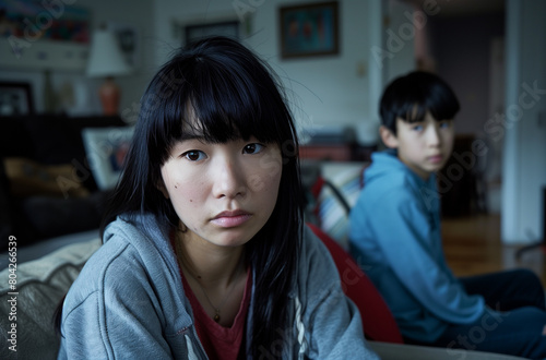 Asian woman with black hair and bangs sitting on the couch in her living room, worried about her teenage son standing behind her.