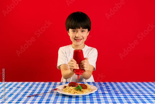 Фотография Smiling Korean boy sitting at table and squeezing ketchup sauce on plate with spaghetti and meatballs against red background