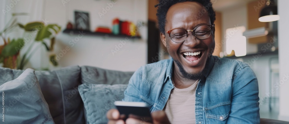 Smiling Black African American man on a sofa in living room having a video call with his friends and family on his smartphone. Smiling Black African American man on a sofa in living room having a video call with his friends and family on his smartphone.