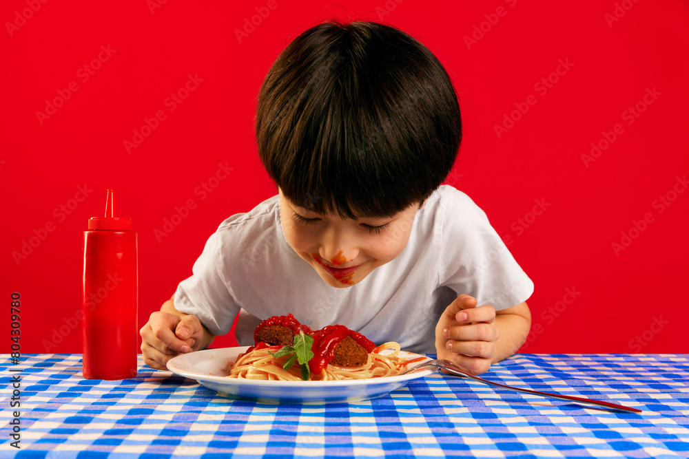 Little boy, child with dirty ketchup face sitting at table and eating delicious spaghetti with meatball and tomato sauce against red background. Food, childhood, emotions, meal, menu, pop art concept Little boy, child with dirty ketchup face sitting at table and eating delicious spaghetti with meatball and tomato sauce against red background. Food, childhood, emotions, meal, menu, pop art concept