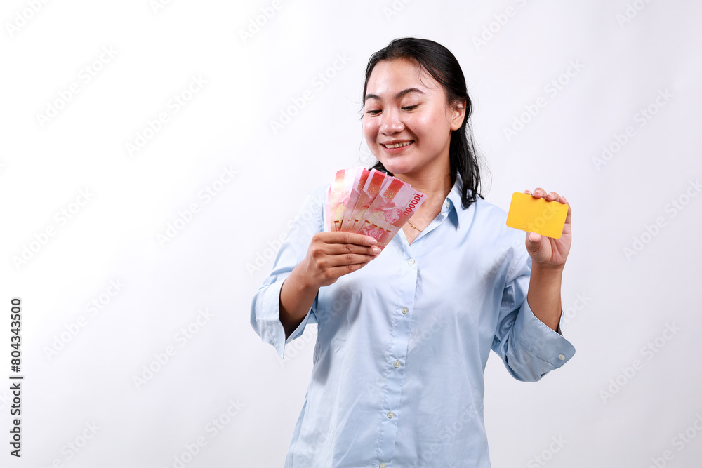 Happy asian woman holding credit card and Indonesian money rupiah, smiling, posing against white studio background