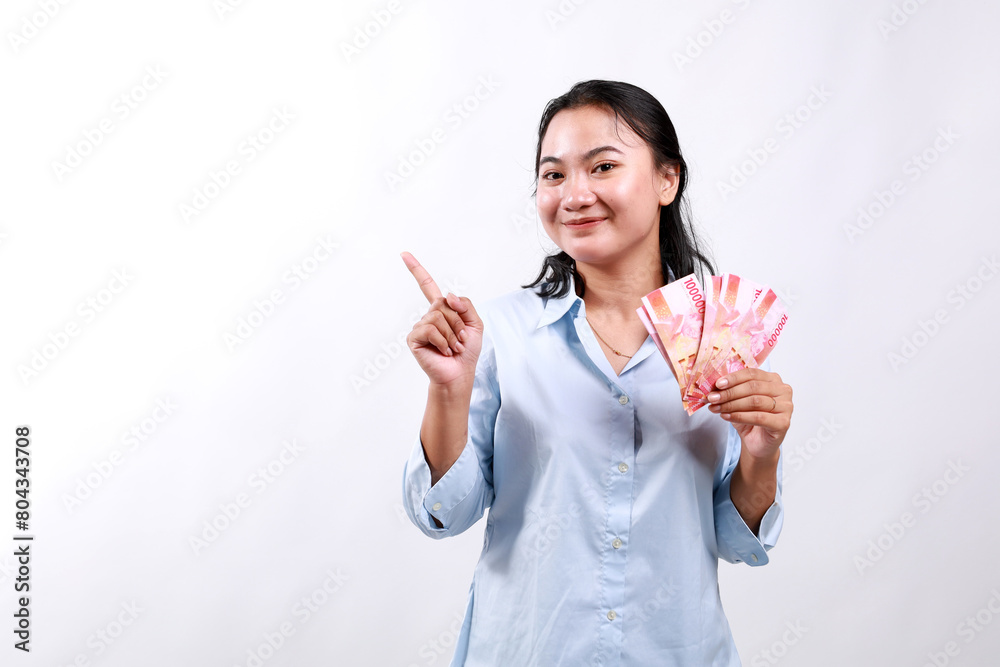 Smiling young modern asian woman, pointing at empty space, holding cash money rupiah, standing over white background looking at the camera Smiling young modern asian woman, pointing at empty space, holding cash money rupiah, standing over white background looking at the camera