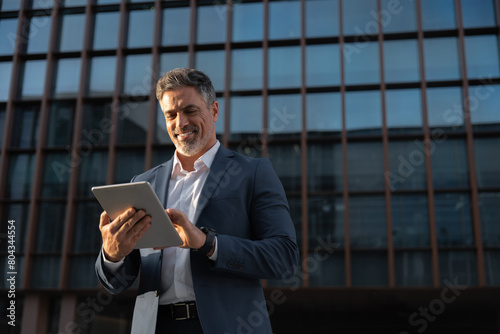Focused successful mature Indian or Latin entrepreneur businessman holding digital pc tablet standing outdoor at business office building. Hispanic smiling man in suit working using touchpad computer photo