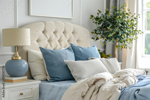 Plant, books and lamp standing on white and gray nightstands in bright and cozy bedroom interior with decorative pillows on bed with blue bedding. Real photo