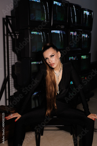  A beautiful girl in a black jumpsuit with an open neckline and high heels, against the backdrop of old working TVs standing in a row. A portrait of girl with bright makeup, winged eyes, big lips.