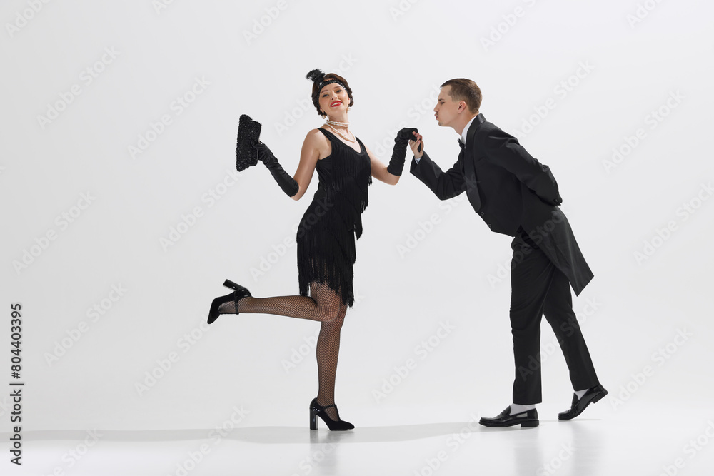 Man in classic black tuxedo and woman in flapper dress performing Charleston dance isolated against white studio background. Concept of art, retro and vintage, hobby, entertainment, 20s Man in classic black tuxedo and woman in flapper dress performing Charleston dance isolated against white studio background. Concept of art, retro and vintage, hobby, entertainment, 20s