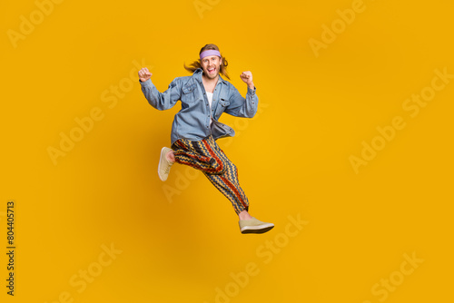 Full size photo of cool young man raise fists jump empty space wear denim shirt isolated on yellow color background