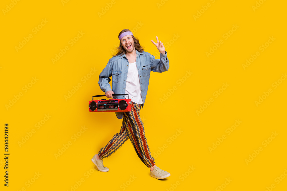 Naklejka premium Full size profile photo of cool young man boombox show v-sign walk empty space wear denim shirt isolated on yellow color background