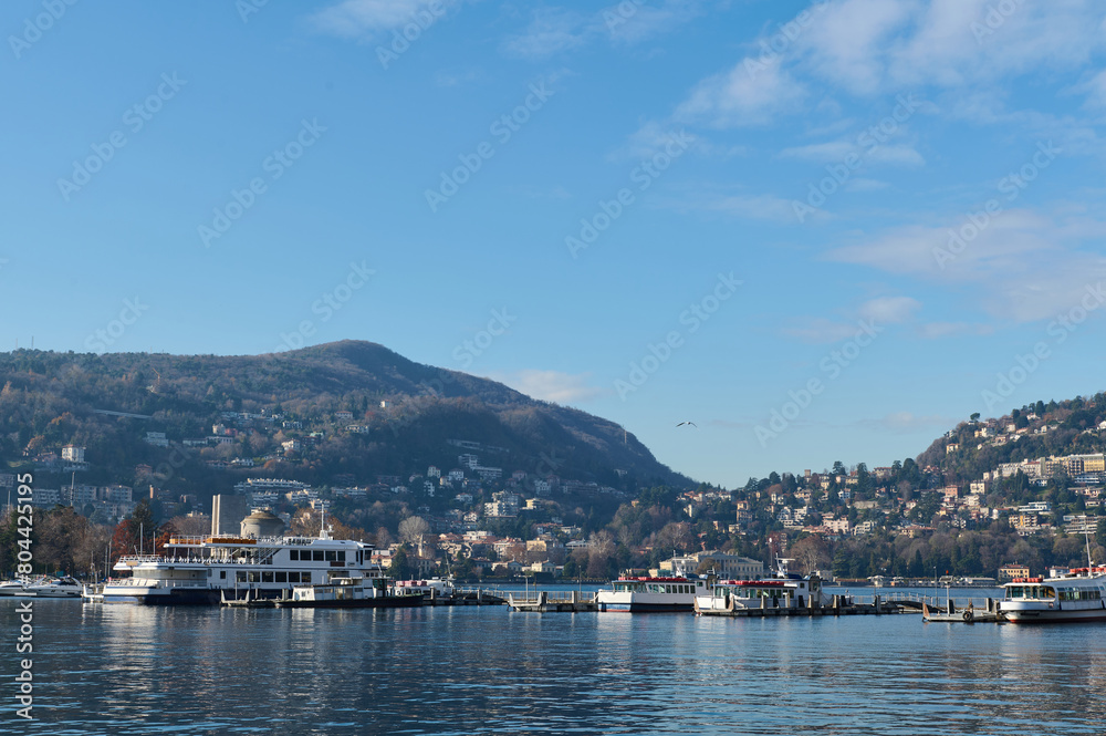 Beautiful view of floating boats and ships reflecting in the clean clear water of the lake of Como, against Italian Alps and blue sky background on a cold sunny winter day. Tourism and travel concept