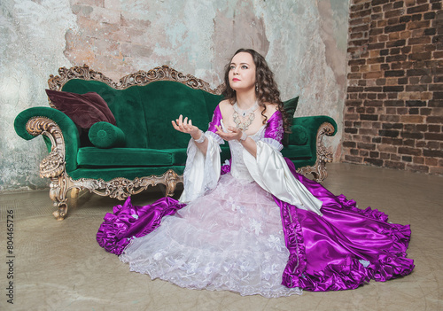 Beautiful woman in fantasy white and purple rococo style medieval dress sitting on the floor near sofa and praying