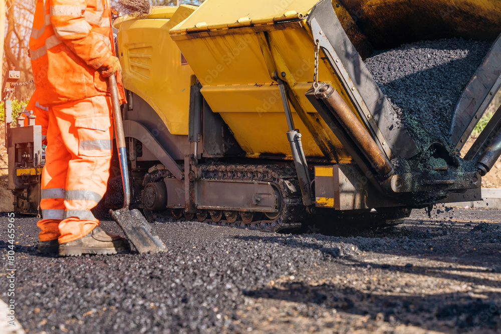 Asphalt paver with hot tarmac roadworker operator in hi-viz laying new road surface Asphalt paver with hot tarmac roadworker operator in hi-viz laying new road surface