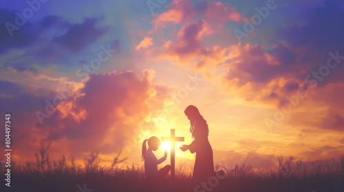 Silhouette of mother and child kneeling in prayer together, side view at sunset. Large cross in background. Mother is standing up while the son sits on his knees holding hands with her praying to God