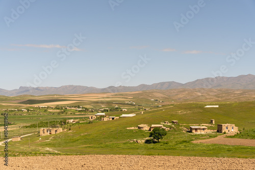 A rural landscape with a few houses and a tree. The sky is blue and there are no clouds