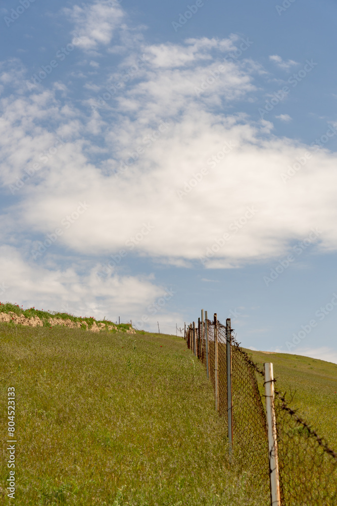 Obraz premium A fence is on a hillside with a clear blue sky. The fence is old and rusted