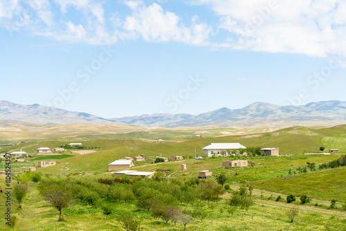 A rural area with a few houses and a few trees. The sky is blue and the sun is shining