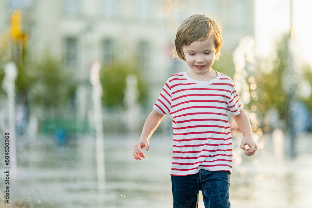 Naklejka premium Cute little boy having fun with water in city fountain. Child playing water games outdoors on hot day. Summer activities for children.