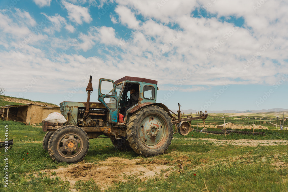 Obraz premium An old blue tractor is parked in a field. The tractor is rusty and has a worn-out appearance. The sky is clear, and the sun is shining brightly. The scene gives off a sense of nostalgia