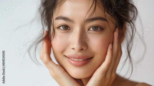 An appealing Asian woman with luminous skin, wearing a smile while smoothly spreading cream on her face. She boasts dark hair and chooses not to wear any makeup. The setting features a white backgroun