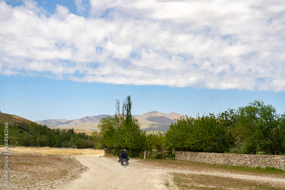 Fototapeta premium A man is riding a motorcycle on a dirt road in the middle of a field. The sky is clear and the sun is shining brightly. The man is wearing a helmet and he is enjoying the ride. The scene is peaceful
