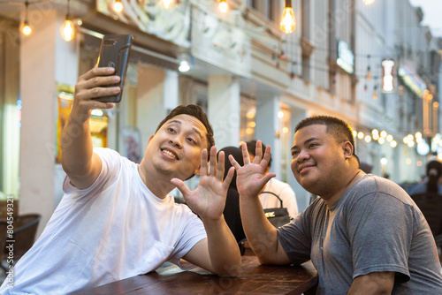 Asian Bestfriends Enjoying Outdoor Cafe, One holding his phone for Taking Selfie or talking in video call with other friend. photo