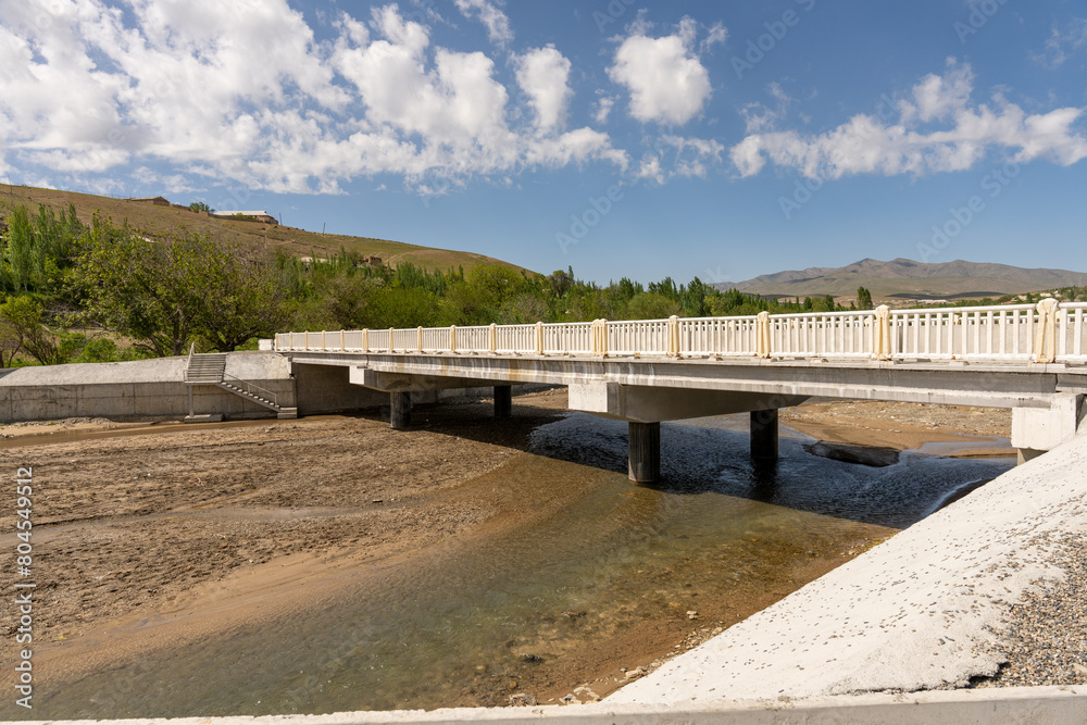 A bridge spans a river with a rocky shoreline. The bridge is made of concrete and has a white railing. The water is calm and the sky is clear A bridge spans a river with a rocky shoreline. The bridge is made of concrete and has a white railing. The water is calm and the sky is clear