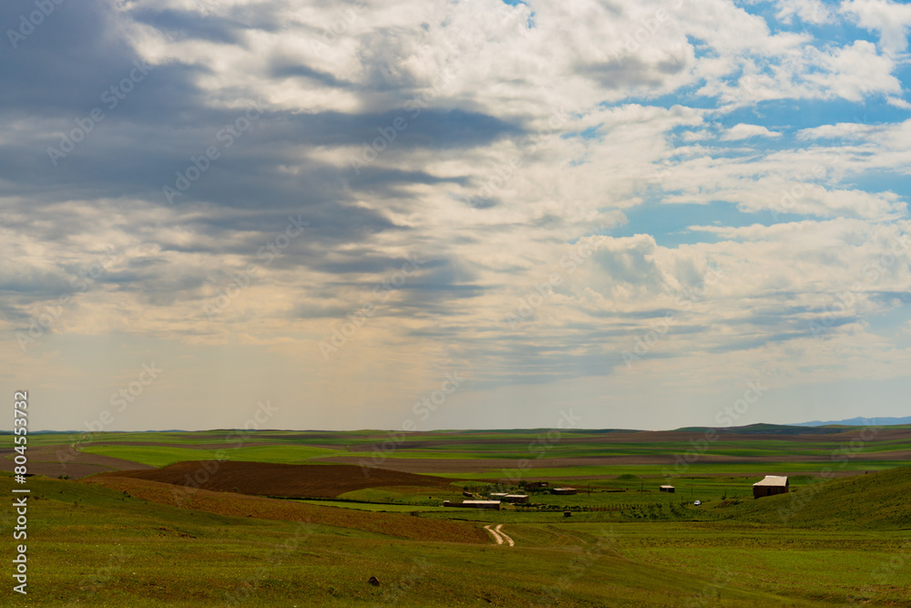 A large, open field with a few buildings in the distance. The sky is cloudy and the sun is shining through the clouds A large, open field with a few buildings in the distance. The sky is cloudy and the sun is shining through the clouds